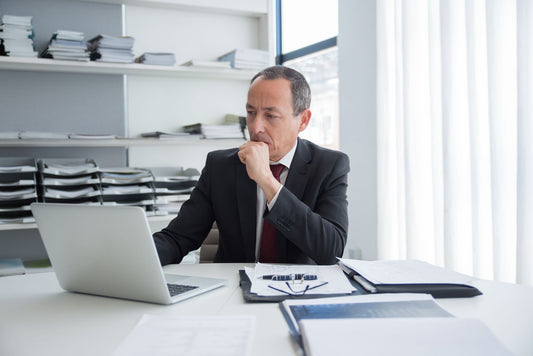 Business owner at his desk looking at a laptop and thinking through his options, with folders and paperwork around him