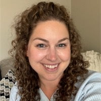 Blonde, curly haired smiling white woman, wearing a blue cardigan in front of a beige background