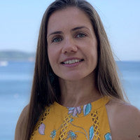 Blonde, white, smiling woman wearing a yellow, floral print top in front of a water landscape
