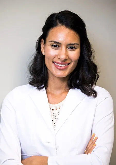 Black haired POC woman wearing a white lab coat and crossing her arms in front of a beige, plain backdrop