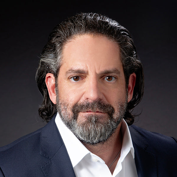 Straight faced white man with a greying beard and black hair. He is in a suit and in front of a grey and black backdrop