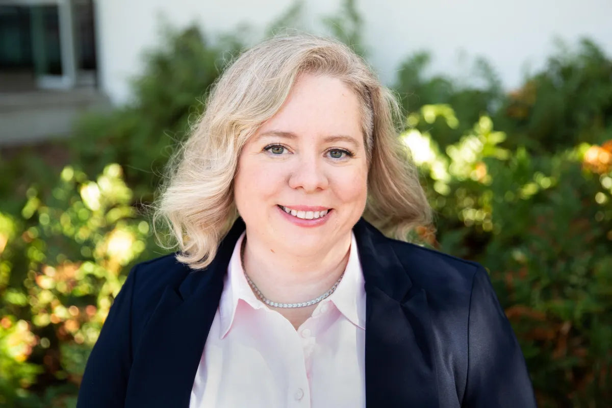 Smiling blonde white woman wearing a pink shirt and navy blue blazer. She is standing in front of blurred out shrubbery 