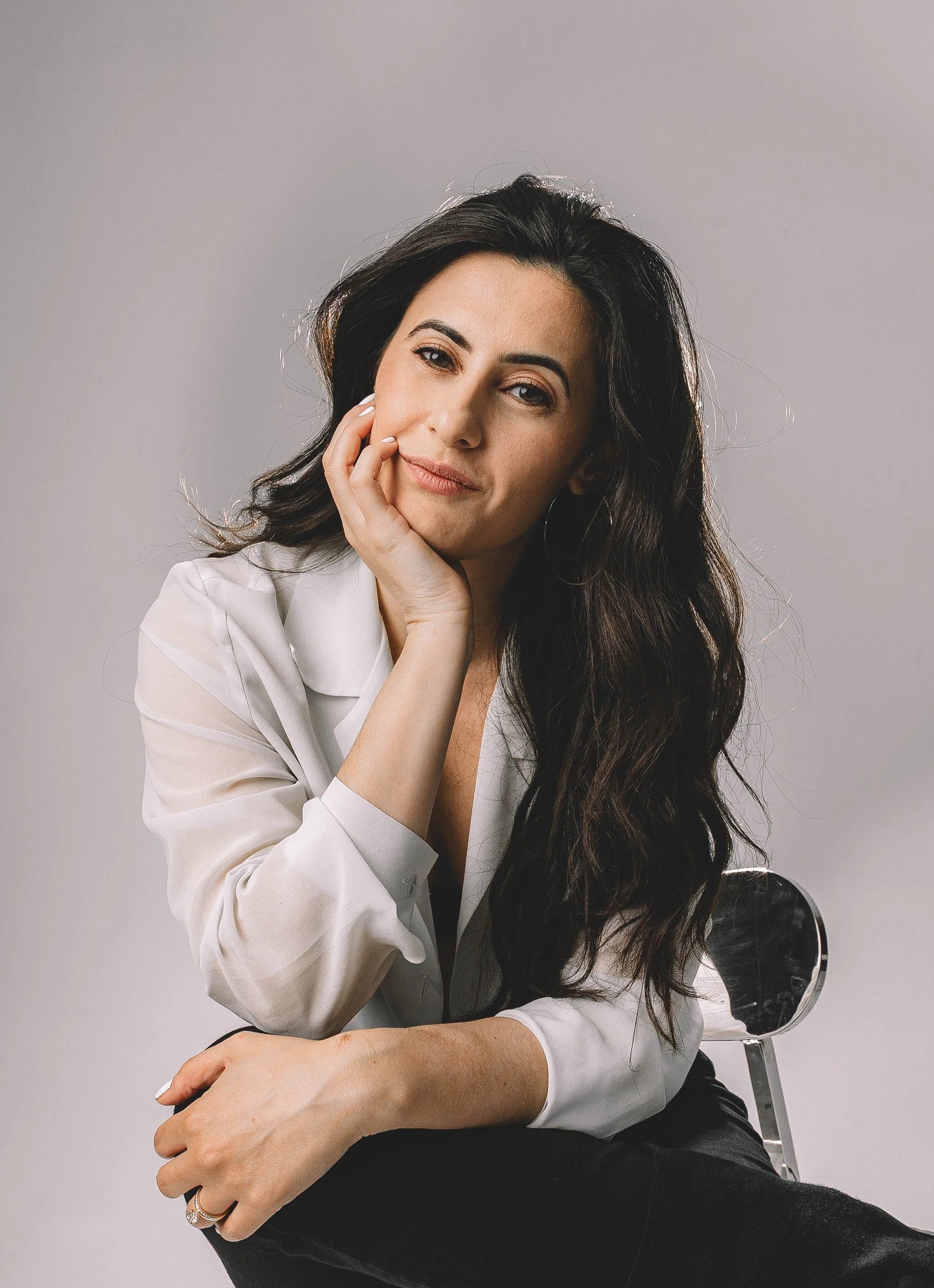 Woman with long, curled black hair, sitting with one leg over the other in a chair with a white backdrop