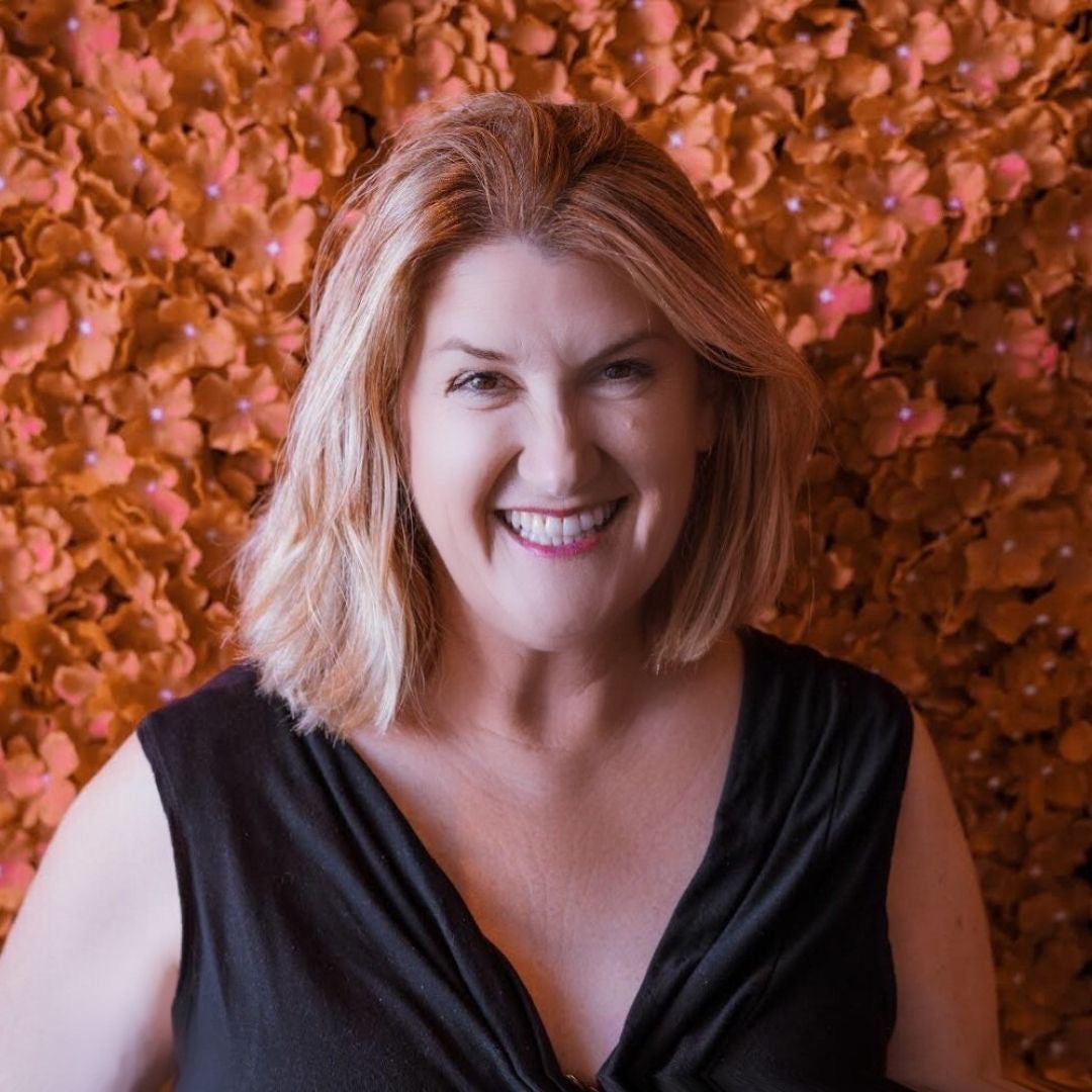 Smiling white, blonde woman with a bob in a black low cut top and in front of a backdrop of orange fake flowers