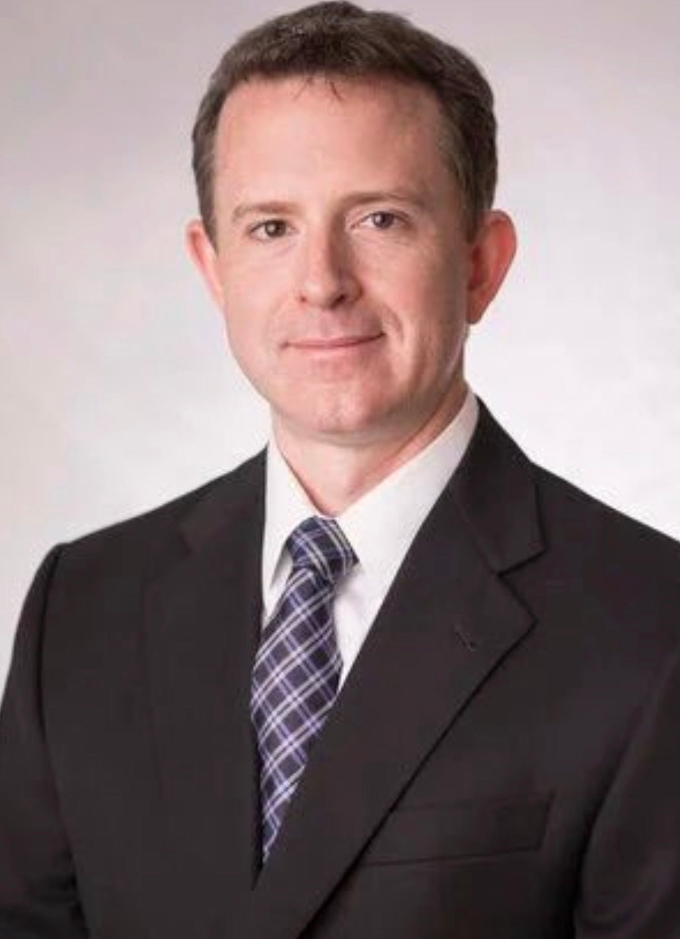 White man with brown short hair, wearing a black suit and blue tie in front of a white background