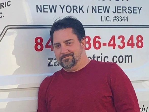 Smiling white man with black hair and a beard, wearing a white shirt and posed in front of his company’s van