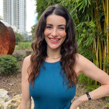 A smiling woman with long wavy brown hair wearing a sleeveless teal-blue top, standing outdoors in front of green bamboo and a city building.
